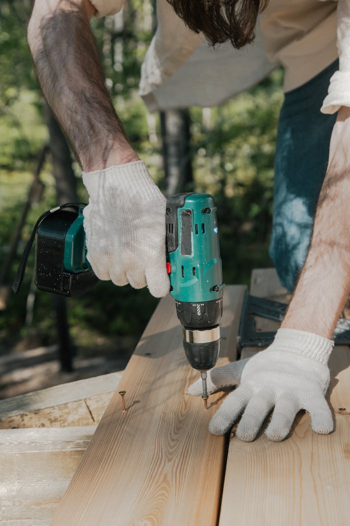 Adult using a power drill on a wooden surface, wearing white gloves, outdoors in daylight.